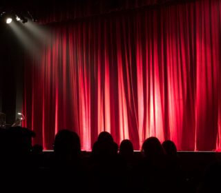 A photo showing a red theatre curtain and members of the audience