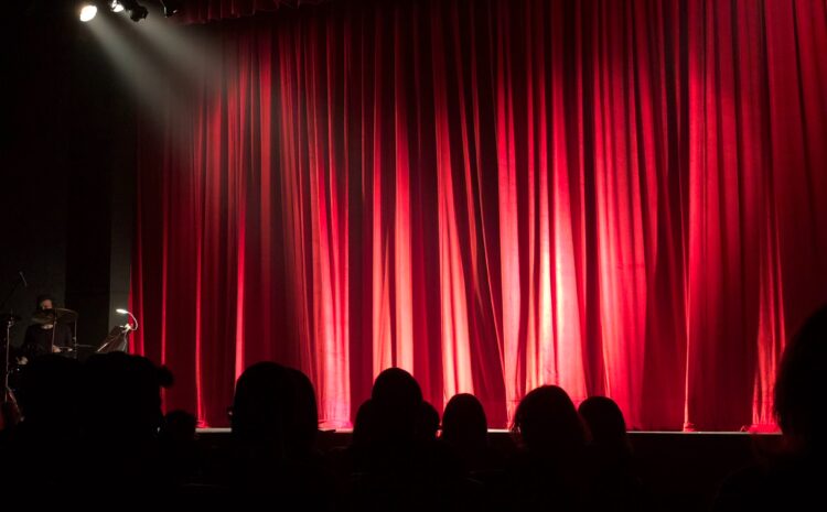 A photo showing a red theatre curtain and members of the audience