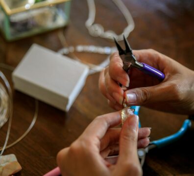 Close-up of hands making jewelry with pliers, holding a wire-wrapped stone pendant on a wooden table with tools, wire, and materials scattered around.