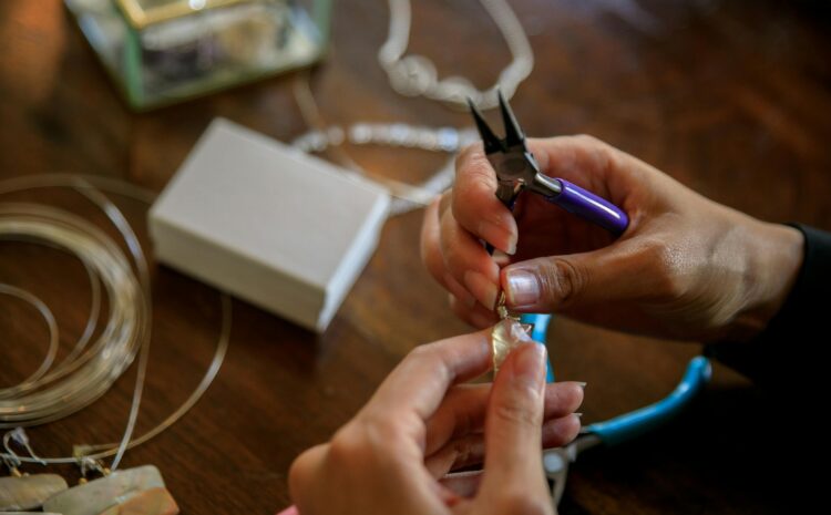 Close-up of hands making jewelry with pliers, holding a wire-wrapped stone pendant on a wooden table with tools, wire, and materials scattered around.