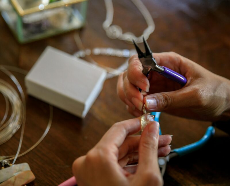 Close-up of hands making jewelry with pliers, holding a wire-wrapped stone pendant on a wooden table with tools, wire, and materials scattered around.