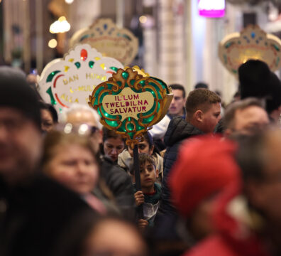 An image showing A crowd of people taking part in the traditional Baby Jesus Procession in Valletta a few days before Christmas. At the center, a young child holds a decorative illuminated sign reading “Illum Twelidna Salvatur,” surrounded by others carrying similar festive signs as they walk through a busy, warmly lit street.