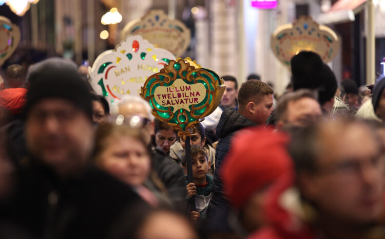 An image showing A crowd of people taking part in the traditional Baby Jesus Procession in Valletta a few days before Christmas. At the center, a young child holds a decorative illuminated sign reading “Illum Twelidna Salvatur,” surrounded by others carrying similar festive signs as they walk through a busy, warmly lit street.
