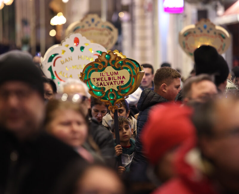 An image showing A crowd of people taking part in the traditional Baby Jesus Procession in Valletta a few days before Christmas. At the center, a young child holds a decorative illuminated sign reading “Illum Twelidna Salvatur,” surrounded by others carrying similar festive signs as they walk through a busy, warmly lit street.
