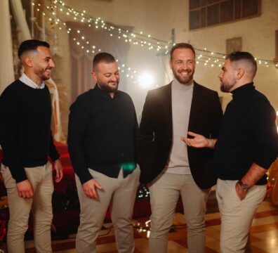 A photo showing the Maltese vocal group Sterjotipi standing together in a warmly lit Christmas-themed setting, smiling and chatting in front of festive lights and decorations