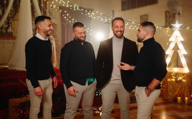 A photo showing the Maltese vocal group Sterjotipi standing together in a warmly lit Christmas-themed setting, smiling and chatting in front of festive lights and decorations