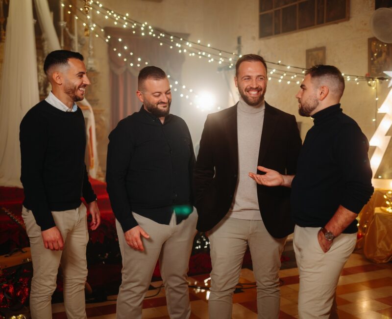 A photo showing the Maltese vocal group Sterjotipi standing together in a warmly lit Christmas-themed setting, smiling and chatting in front of festive lights and decorations