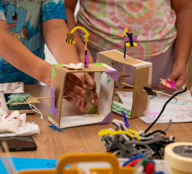 Children’s hands building small cardboard tinkering projects decorated with colorful paper bees and butterflies, surrounded by craft supplies on a wooden table.