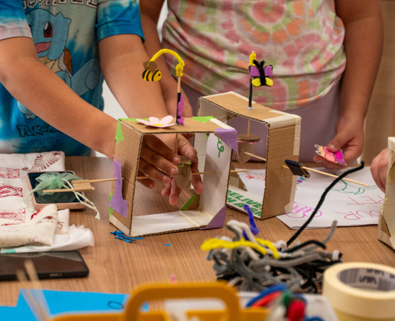 Children’s hands building small cardboard tinkering projects decorated with colorful paper bees and butterflies, surrounded by craft supplies on a wooden table.